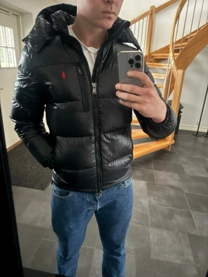 Svart pufferjacka från Polo Ralph Lauren - Säljer en svart pufferjacka från Polo Ralph Lauren med röd logga på bröstet. Jackan har huva, dragkedja framtill och en extra bröstficka med zip. Materialet är glansigt. Perfekt för kalla dagar. Inga skador. Köpt från Ralph lauren. 