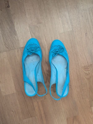 Turkosa ballerinaskor i skinn, strl 39 - Snygga turkosa ballerinaskor i skinn med öppen häl och dekorativ rosett framtill. Skorna har rund tå, små håldetaljer under skon och låg klack i trälook. Perfekta för dig som vill sticka ut med färg och stil..den blå färgen är inte sliten