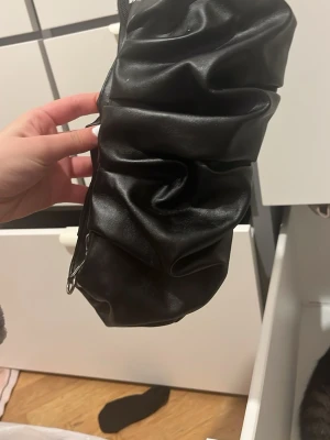 Svart axelväska i syntet med rynk - Säljer en svart axelväska i syntetmaterial med cool rynkad design. Väskan har dragkedja upptill och metallfästen på sidorna. Perfekt storlek för det viktigaste och ger en trendig touch till din outfit.