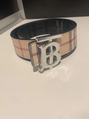 Rutig och svart bälte med TB-spänne - Säljer ett Burrberry bälte med klassiskt rutigt mönster i beige, svart och rött samt en sida i svart med präglat rutmönster. Stort silvrigt TB-spänne framtill. Perfekt accessoar för att lyfta din outfit.