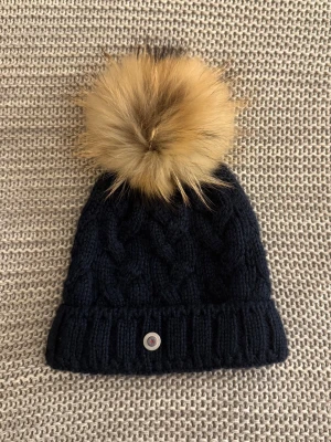 Marinblå Bogner Stickad Mössa med Pälsboll - En elegant marinblå mössa med en stor, lyxig pälsboll från Bogner. ✨ Köpt på NK. Perfekt skick! Den ser ut som ny, då den endast har använts 1-2 gånger. Mössan är stickad i ett klassiskt mönster, och är varm, mjuk och stretchig. Den har en uppvikt kant, samt en liten Bogner-logga i metall framtill. Insidan är i ett mjukt ’fleece’-material. Passar utmärkt för att hålla värmen under kyligare dagar! Pälsen: Asiatic Raccoon, ursprung: Finland
