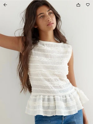 Vit volangblus med struktur - Supersöt vit blus med volangkant nertill och strukturerat tyg i horisontella ränder. Ärmlös modell med rund halsringning, perfekt till jeans eller kjol för en fräsch look.