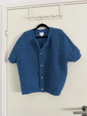 Blå stickad kofta med kort ärm - Kortärmad blå stickad kofta med strukturmönster och fyra dekorativa knappar framtill. Koftan har en avslappnad passform och ribbade kanter vid hals, ärmslut och nederkant. Perfekt för lager på lager under vår och höst. Jag har vanligtvis xs och denna passade jätte bra som en oversized tröja (den är strl XL) använd en gång