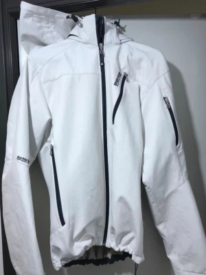 Vit vindjacka  - Säljer en vit vindjacka med svart dragkedja och flera fickor. Jackan har huva med snörning och lång ärm. Perfekt för blåsiga dagar och har en sportig look. Vind & regn jacka 