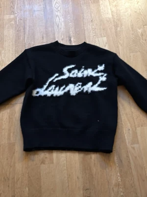 Saint Laurent stickad tröja M - Säljer en svart stickad tröja från Saint Laurent i storlek M. Tröjan har ett stort vitt Saint Laurent-tryck framtill i fluffig stil och ribbade muddar vid ärmslut och nederkant. Perfekt för dig som vill ha en statement piece i garderoben.