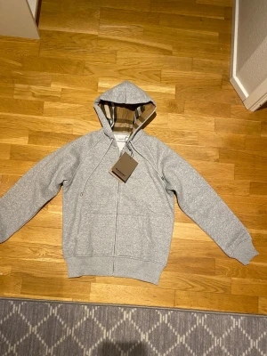 Burberry Zip-up Hoodie Helt Ny!!! -  Upplev lyx och stil med denna exklusiva zip-up hoodie från Burberry. Tillverkad i mjuk, ljusgrå bomull med en bekväm passform i storlek M. Insidan av huvan pryds av Burberrys ikoniska rutmönster i beige, svart och vitt – en subtil men distinkt detalj som utstrålar elegans. Helt Ny!!!