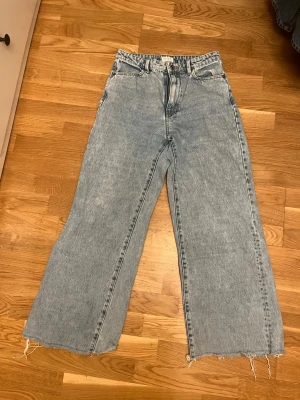 Wide jeans från Lindex, ljusblå - Säljer ett par ljusblå jeans från Lindex i modell med breda ben och råa, fransiga benslut. Klassisk femficksdesign och hög midja. Perfekta för dig som gillar en avslappnad och trendig look.