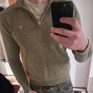 Olivgrön stickad tröja från Morris - Säljer en olivgrön stickad tröja från Morris med halv dragkedja och ribbade muddar. Tröjan har en liten vit broderad logga på bröstet och är långärmad. Perfekt för dig som gillar klassisk men avslappnad stil.