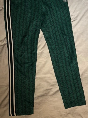   Lacoste Byxor - Säljer ett par gröna träningsbyxor från Lacoste med vita samt svarta ränder längs sidorna. Byxorna är i mycket bra skick, hör av dig vid minsta fundering. Priset är inte hugget i sten🙌