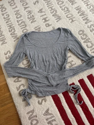 Grå ribbad långärmad topp med snörning - Säljer en grå långärmad topp i ribbat material med snörning i sidorna. Toppen har en rundad urringning och är i mjuk stretchig kvalitet. Perfekt till jeans eller kjol för en chill look.