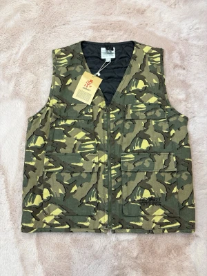 Camouflageväst från Gramicci Outdoor - Säljer en grön och gul camouflageväst från Gramicci Outdoor. Västen har flera stora fickor framtill, dragkedja och cool utility-vibe. Tillverkad i slitstark bomull och perfekt för dig som gillar streetwear eller outdoor-stil.