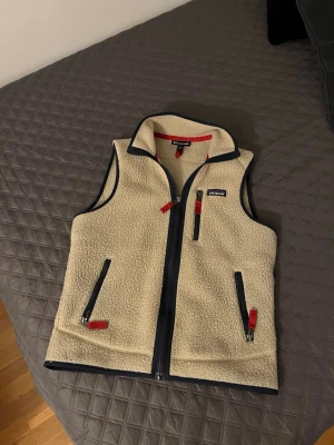 Beige fleeceväst från Patagonia XS - Säljer en beige fleeceväst från Patagonia i storlek XS. Västen har marinblå detaljer, röda dragkedjor och tre fickor framtill. Mjuk och fluffig känsla, perfekt för lager på lager. Klassisk Patagonia-logga på bröstet och hög krage. Hör av dig vid minsta lilla, lägg gärna ett prisförslag🤩🙌🏼