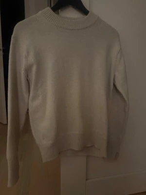 Beige stickad tröja från H&M - En enkel och stilren beige stickad tröja från H&M i storlek S. Tröjan har rund halsringning med bred ribbad kant och långa ärmar med ribbade muddar. Perfekt för lager på lager under kyliga dagar.