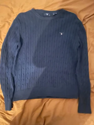 Mörkblå kabelstickad tröja från GANT - Säljer en mörkblå kabelstickad tröja från GANT med rund hals och diskret logga på bröstet. Tröjan har långa ärmar och ribbade muddar vid ärmslut och nederkant. Perfekt för dig som gillar klassisk och stilren look.