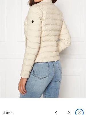 Beige pufferjacka från Rockandblue - Snygg beige pufferjacka från Rockandblue med guldfärgade dragkedjor och diskret logga på ärmen. Jackan har quiltad design, hög krage och två fickor med dragkedja. Perfekt för dig som vill ha en lätt och stilren jacka till vardags.