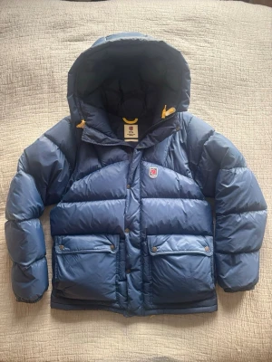 Blå Fjällräven Expedition dunjacka L - Säljer en blå Fjällräven Expedition Series dunjacka i storlek L. Jackan har en stor huva, två rejäla framfickor med knappar och dragkedja framtill. Klassisk Fjällräven-logga på bröstet och gula detaljer vid huvan. Den är i mycket bra skick och har inga defekter.