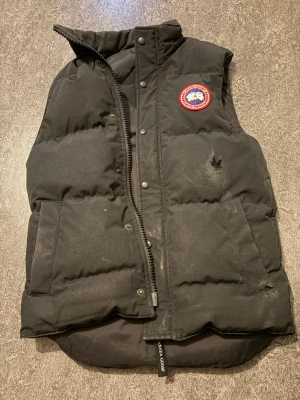Svart dunväst från Canada Goose XS (äkta) - Säljer en svart dunväst från Canada Goose i storlek XS. Västen har hög krage, två stora fickor framtill och klassisk Canada Goose-logga på bröstet. Stängs med dragkedja och tryckknappar. Perfekt för kalla, blåsiga dagar.