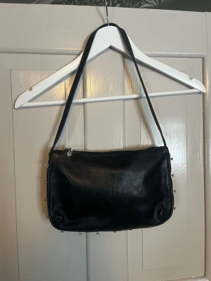 Roberto Cavalli Y2K väska  - Roberto Cavalli axelväska – äkta läder – Y2K  Snygg och tidlös axelväska från Roberto Cavalli i svart mjukt läder. Karaktäristisk Cavalli-design med dekorativa metallnitar runt kanterna. Perfekt storlek för vardag eller kväll, rymmer det nödvändigaste.  • Äkta läder • Justerbar axelrem • Dragkedja upptill • Färg: svart • Original Cavalli-detaljer i metall  Skick: Begagnat i gott bruksskick med normalt slitage och fin patina (se bilder).