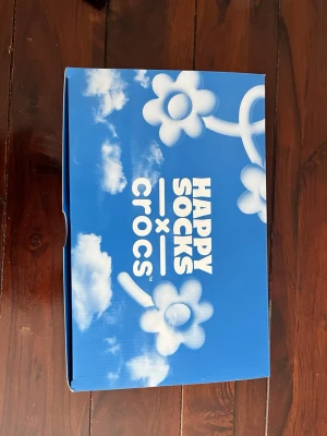 Happy Socks x Crocs blå molnmönstrade - Unika Crocs i samarbete med Happy Socks, blå med vita moln och färgglada pins. Insidan är fodrad med mjuk, fluffig textil i blått och vitt. Perfekta för dig som vill sticka ut och ha det bekvämt. Säljs i originalkartong.