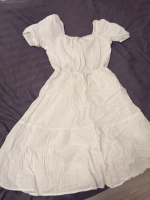 Vit sommarklänning från Lindex - Supersöt vit klänning från Lindex med korta puffärmar och volangkjol. Klänningen har en öppen rygg med knyt och är gjord i ett lätt, strukturerat tyg som känns perfekt för varma dagar. Använd en gång. Köpt för 499kr för några år sen.