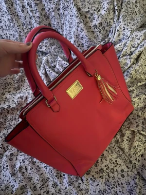 Rosa handväska med tofs och guldskylt - Säljer en snygg rosa handväska från Love med guldfärgad skylt framtill och dekorativ tofs. Väskan har två rejäla handtag, dragkedja upptill och rymligt fack inuti. Perfekt för dig som vill ha en färgklick till din outfit.