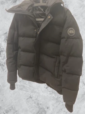 Svart pufferjacka från Canada Goose - Säljer en svart pufferjacka från Canada Goose med huva och klassisk logga på ärmen. Jackan har dragkedja framtill, guldiga knappar och är vadderad för extra värme. Perfekt för kalla dagar och har en clean, stilren look.