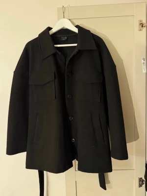 Kappa - Gina Tricot - Säljer en svart kappa från Gina Tricot i ullmix. Jackan har stora bröstfickor, knappar framtill och ett bälte i midjan för extra snygg passform. Perfekt oversized look och långärmad modell som passar till lager på lager.