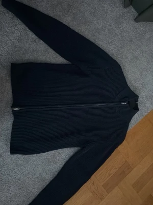 Mörkblå stickad kofta från 157 - Säljer en mörkblå stickad kofta från 157 med grov ribbad struktur och hel dragkedja framtill. Tröjan har hög krage och långa ärmar, perfekt för kyliga dagar. Enkel och stilren design som funkar till det mesta.