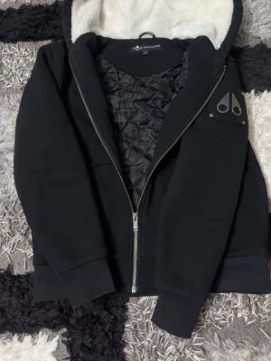 Svart hoodiejacka från Moose Knuckles - Svart hoodiejacka från Moose Knuckles med dragkedja, quiltat innerfoder och vit fuskpäls i huvan. Jackan har en ikonisk logga i metall på ärmen och stora fickor framtill. Perfekt för dig som vill ha en stilren och varm jacka med streetkänsla.