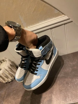 Nike Air Jordan 1 High UNC - Säljer ett par Nike Air Jordan 1 High i UNC-färgway. Skorna har vit bas med ljusblå och svarta detaljer, svart swoosh och svarta skosnören. Klassisk hög siluett med perforerad tåbox och ikoniska Jordan-loggan på sidan. Tillverkade i skinn. Äkta ( scanna QR-koden så kommer du till Nike.com). Använda bara en gånger på sommaren så inte varit i snö, slask eller liknande. Skicka bara vid frågor 