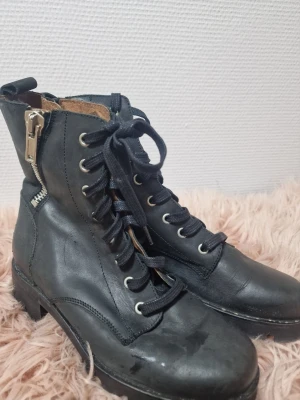 Svarta kängor med snörning och dragkedja - Tuffa svarta kängor i skinn med snörning framtill och dragkedja på sidan. Grov sula i syntet och rund tå ger en cool look. Tillverkade i Spanien och fodrade med läder för extra komfort. Perfekta för dig som vill ha en edgy stil.