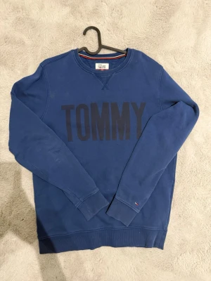 Blå sweatshirt från Tommy Hilfiger - Säljer en blå sweatshirt från Tommy Hilfiger Denim med stort svart 'TOMMY'-tryck på bröstet och klassisk rund hals. Tröjan har långa ärmar, ribbade muddar och liten flaggdetalj vid ärmslutet. Perfekt för dig som gillar stilren och enkel design.