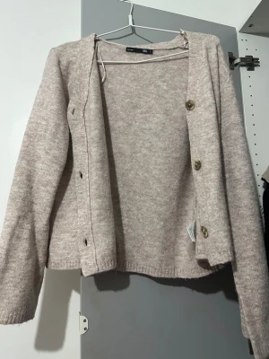 Beige kofta med knappar från 157 - Mysig beige kofta från 157 med v-ringad hals och stora knappar framtill. Koftan är stickad i mjukt material och har långa ärmar samt ribbade muddar vid ärmslut och nederkant. Perfekt för lager på lager under kyliga dagar.