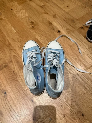Ljusblå Converse All Star sneakers - Säljer ett par klassiska Converse All Star high tops i ljusblått tyg med vita gummidetaljer och vita snören. Skorna har rund tå, platt sula och den ikoniska loggan på sidan. Perfekta för dig som vill ha en retro och avslappnad stil.