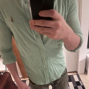 Ljusgrön skjorta från Polo Ralph Lauren - Säljer en ljusgrön skjorta från Polo Ralph Lauren i klassisk oxfordstil. Skjortan har långa ärmar, knappar framtill och den ikoniska broderade loggan på bröstet. Tillverkad i mjuk bomull och passar perfekt till jeans eller chinos.