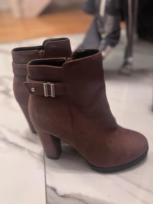 Bruna mocka stövletter med klack - Snygga bruna stövletter i mocka med bred klack och rund tå. Skorna har dragkedja på insidan och ett dekorativt spänne med metall-detalj på sidan. Perfekta för höst och vinter och ger en stilren look till din outfit.