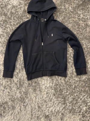 Polo Ralph Lauren hoodie Marinblå zip-hoodie - Polo Ralph Lauren hoodie (zip-up) Marinblå zip-hoodie från Polo Ralph Lauren med vit broderad logga på bröstet. Klassisk modell med huva, dragkedja och fickor framtill. Mjuk och bekväm – passar både till vardags och casual outfits.