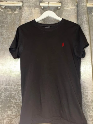 Svart t-shirt från Ralph Lauren - Klassisk svart t-shirt från Ralph Lauren med den ikoniska röda logon broderad på bröstet. T-shirten har rund hals och korta ärmar, perfekt basic-plagg till garderoben. Tillverkad i mjuk bomull för skön känsla hela dagen.