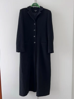 Svart lång kappa från Kookai - Säljer en svart lång ullkappa från Kookai i klassisk rak modell med krage och fyra knappar framtill. Kappan har diskreta fickor och är helfodrad, perfekt för kyliga dagar. Stilsäker och enkel att matcha med olika outfits.