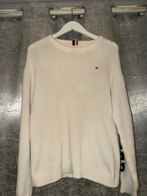 Vit stickad tröja Tommy Hilfiger - Säljer en vit stickad tröja från Tommy Hilfiger med rund halsringning och lång ärm. På ena ärmen står det 'HILFIGER' i stora bokstäver och på bröstet finns den klassiska lilla loggan. Perfekt för dig som gillar stilrena och sportiga plagg.