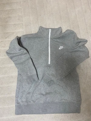 Grå Nike half zip hoodie M - Säljer en grå hoodie från Nike i storlek M. Tröjan har en vit half zip-dragkedja framtill, hög krage och klassisk Nike-logga på bröstet. Tillverkad i mjuk bomullsblandning med långärmad passform och ribbade muddar. Perfekt för chill dagar eller träning. 100% äkta från Zalando.