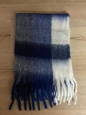 Rutig fluffig väst i blå och vit - Säljer en supermjuk och fluffig väst med rutigt mönster i marinblått, vitt och grått. Västen har långa fransar nedtill och känns riktigt cozy. Perfekt för dig som vill ha en statement piece i garderoben.