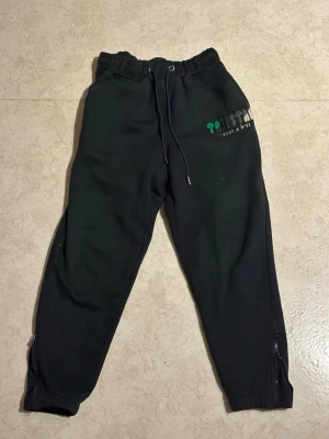 Svarta Trapstar  mjukisbyxor - Svarta mjukisbyxor med resår i midjan och snörning framtill. Byxorna har tryck på ena benet och dragkedjor vid bensluten för extra stil. Tillverkade i mjukt bomullsmaterial som är skönt att ha på sig. Perfekta för chill dagar eller när du vill vara bekväm. Vill bara få sålt, tveka inte vid frågor eller att buda lägre pris❗️