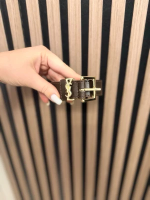 Brunt YSL bälte med gulddetaljer - Snyggt brunt bälte från YSL med klassiskt guldspänne och YSL-logga i metall. Bältet är gjort i skinn och har en stilren, tidlös design som passar till många outfits.