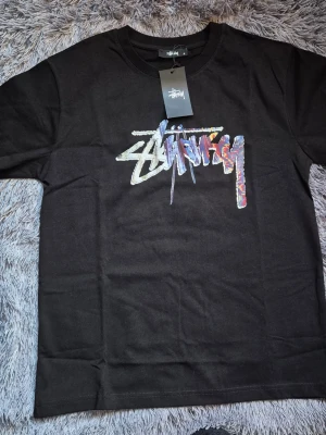 Svart Stüssy t-shirt med tärningsprint - Svart t-shirt från Stüssy med färgstarkt logotryck på bröstet och stor grafik med tärningar på ryggen. Klassisk passform och rund hals. Tillverkad i mjuk bomull som känns skön mot huden. Perfekt för dig som gillar streetwear och vill sticka ut.  