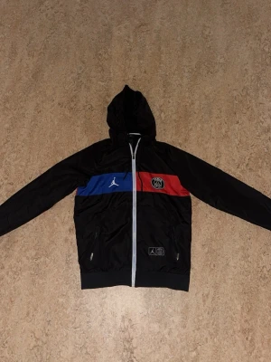 PSG  - Säljer en fet PSG x Jordan vindjacka i svart med blå och röd rand över bröstet. Jackan har huva, hel dragkedja, två fickor med dragkedja och stora PARIS-trycket på ryggen. Tillverkad i lätt polyester, perfekt för streetwear.