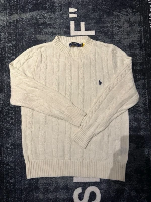 Vit kabelstickad tröja Polo Ralph Lauren - Kabelstickad vit tröja från Polo Ralph Lauren med klassisk rund hals och broderad marinblå logga på bröstet. Tröjan har ribbade muddar och är långärmad, perfekt för lager på lager under kyliga dagar.