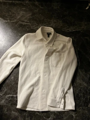 Vit overshirt från H&M  - Säljer en stilren vit overshirt från H&M med klassisk krage, bröstficka och tryckknappar framtill. Skjortan är långärmad och gjord i mjuk bomull som känns skön mot huden. Perfekt att slänga över en t-shirt för en clean look
