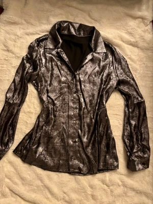 Glittrig silver skjorta med krage - Snygg glittrig skjorta i silver med klassisk krage och långa ärmar. Skjortan har en figurnära passform och knappar framtill samt vid ärmsluten. Materialet är glansigt och ger en riktigt cool look.