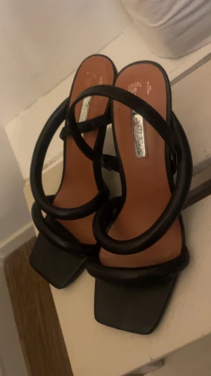 Svarta klackade sandaler från Primark - Säljer ett par svarta sandaler från Primark med höga klackar och fyrkantig tå. Skorna har breda, vadderade remmar i syntetmaterial som korsar över foten för en trendig look. Perfekta för dig som vill ha en edgy vibe till outfiten.